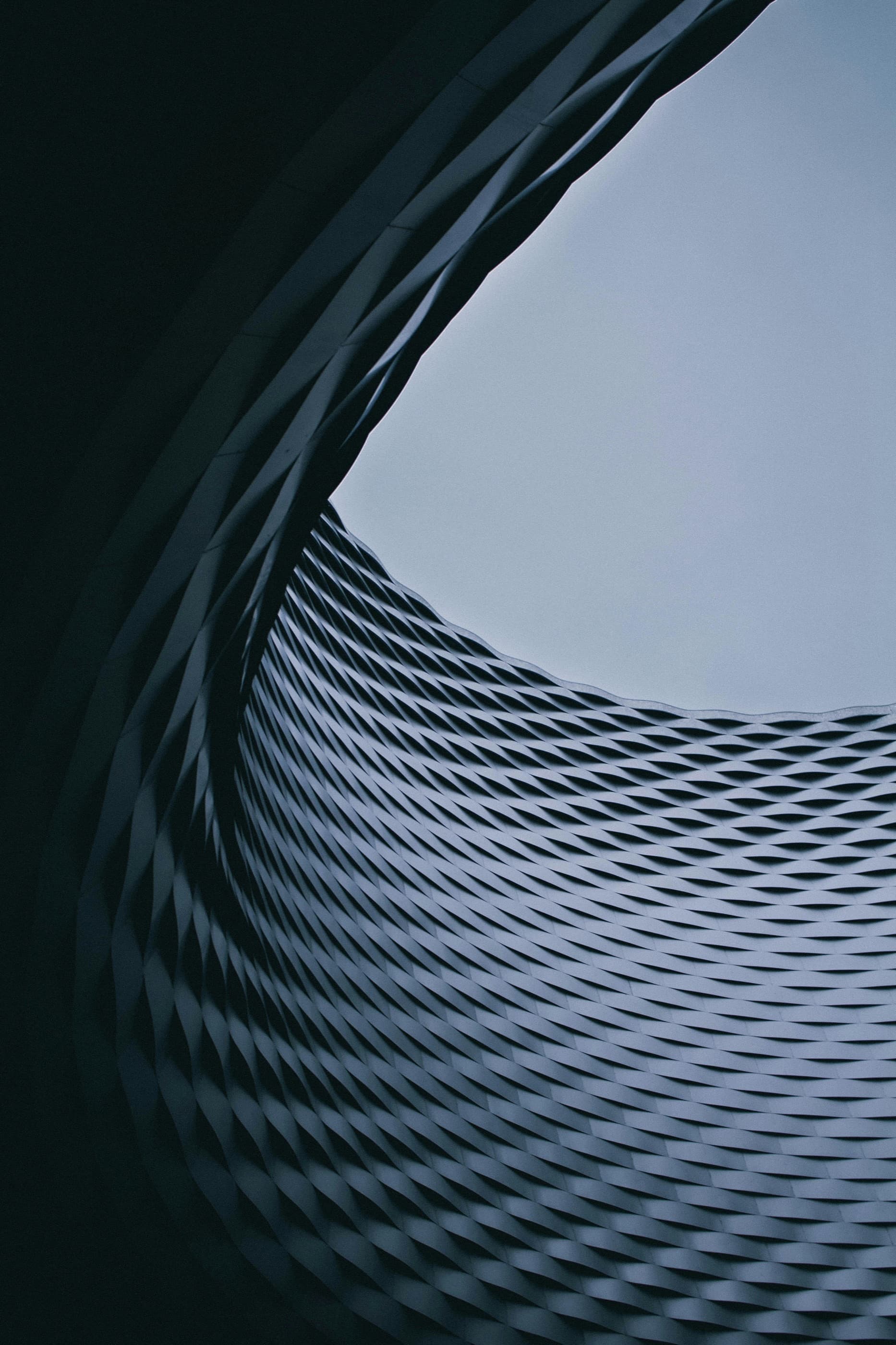 Looking up through a circular void in a woven metal façade to a pale sky