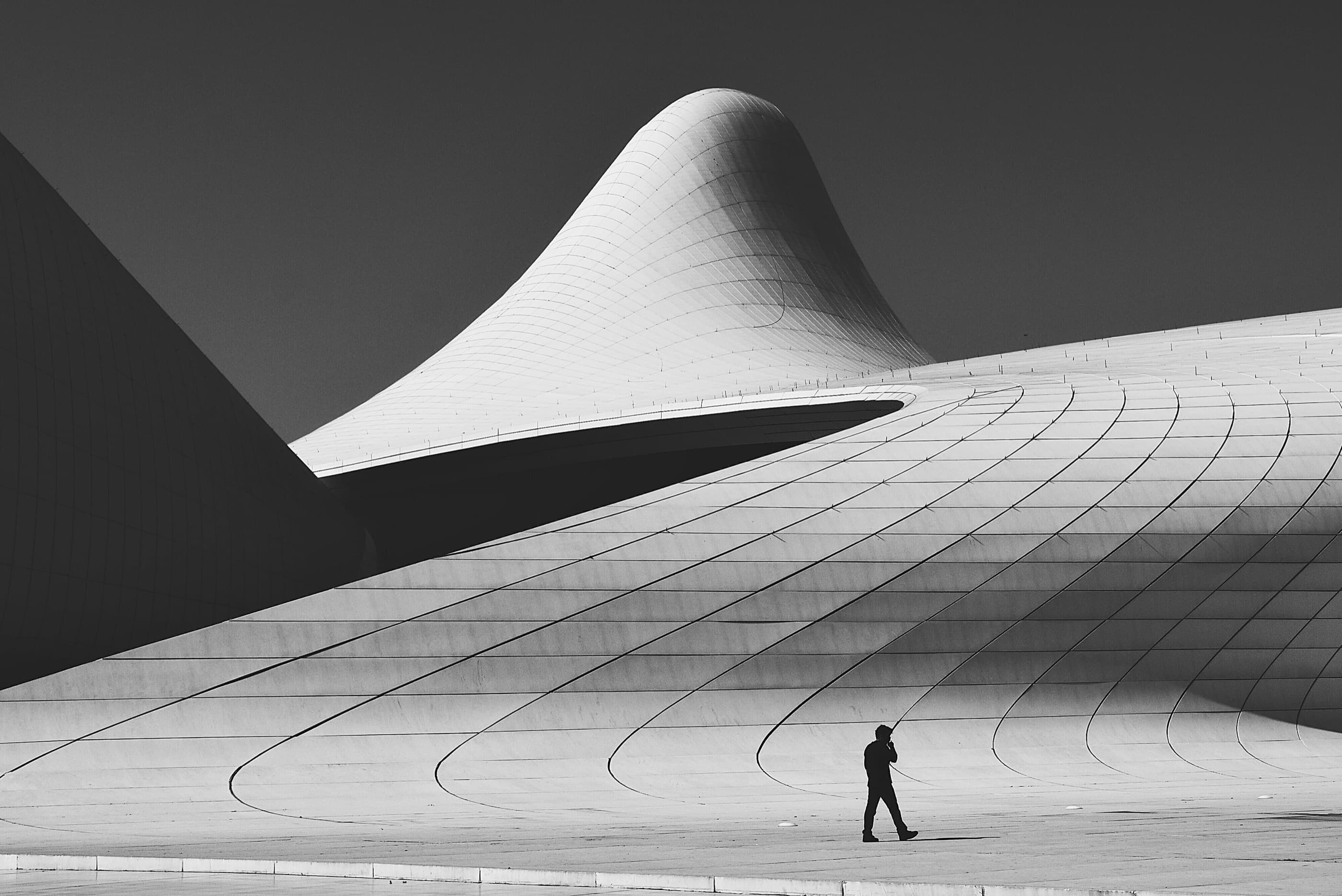 A sweeping white curved concrete building with a lone figure crossing the stepped plaza in front of it